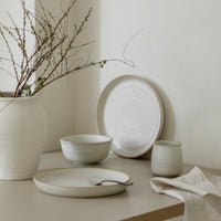 A set of white stoneware dinnerware with a swirl pattern, including a round plate and a bowl, displayed on a neutral-toned surface with a vase of greenery in the background.