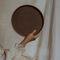A person holding up a round, brandy red ceramic plate against a neutral background.